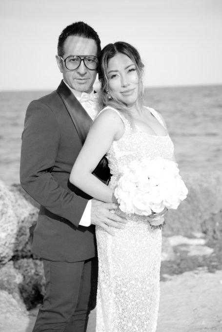 A couple poses together on a beach, the bride holding a bouquet and wearing a sparkling gown.