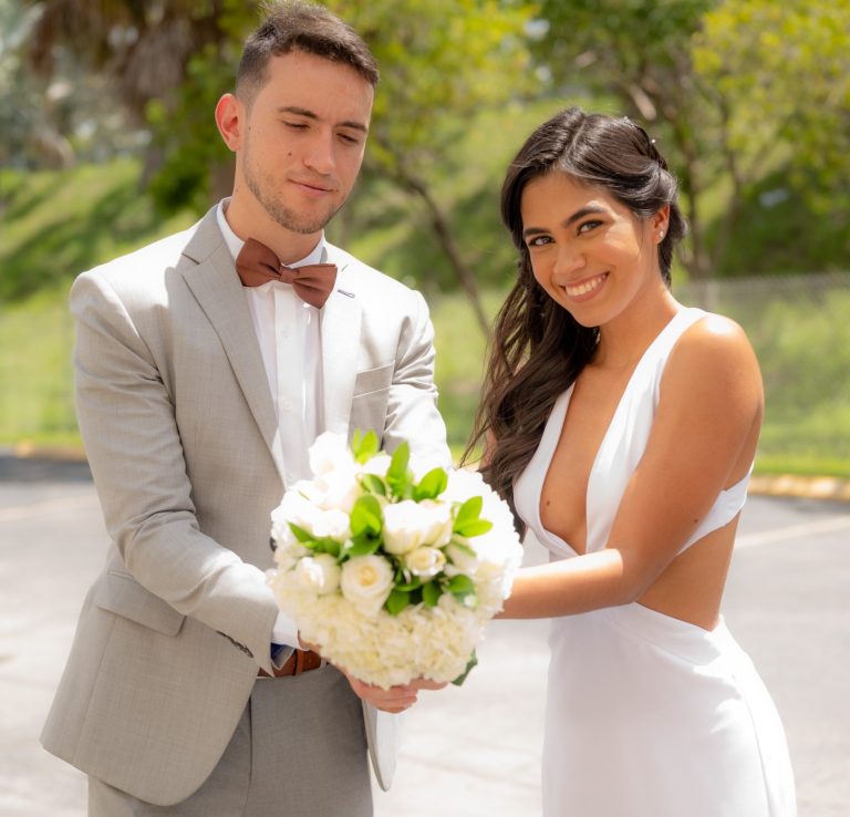 A groom in Miami and his brides holds a bouquet of flowers, smiling outdoors.