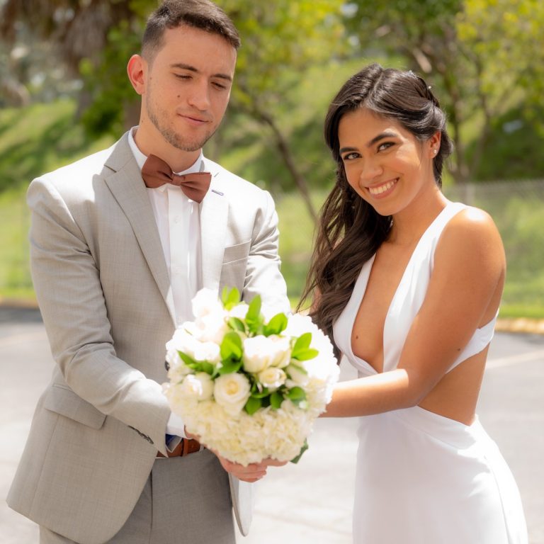 A groom in Miami and his brides holds a bouquet of flowers, smiling outdoors.