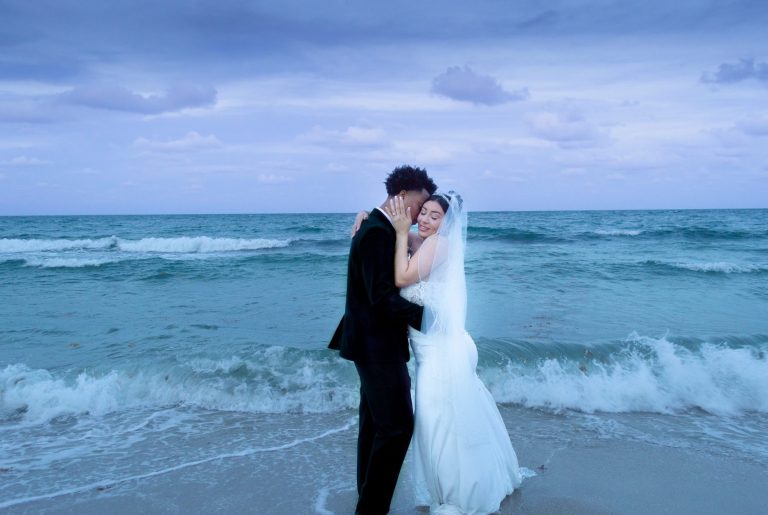 Bride and groom kiss on a beach at sunset, waves gently lapping at their feet.