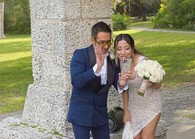 A groom in a blue tuxedo and a bride in a white dress pose for a selfie.