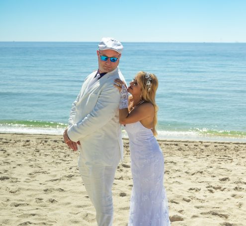 Couple in wedding attire on a beach, smiling and enjoying the sunny day.