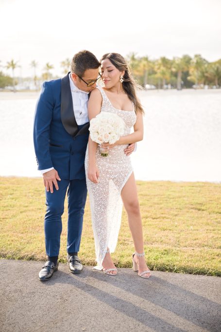 A couple poses romantically outdoors, the woman wearing a sheer, elegant dress and holding flowers.