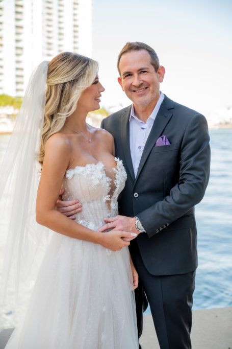Bride and groom smiling together, dressed elegantly by a waterfront.