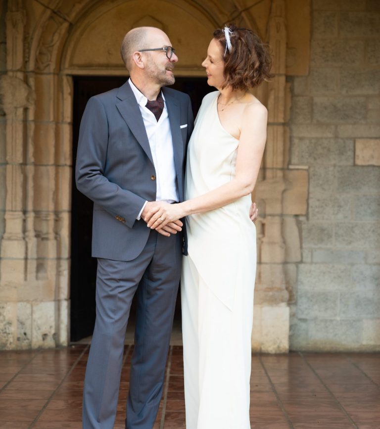 A couple joyfully holding hands, smiling at the camera against a colorful backdrop by Iris Ramil Wedding Officiant.