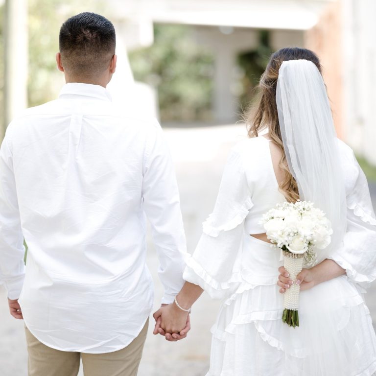 Just married by Iris Ramil wedding officiant, a Bride & groom holds hands, bride with bouquet, walking away.