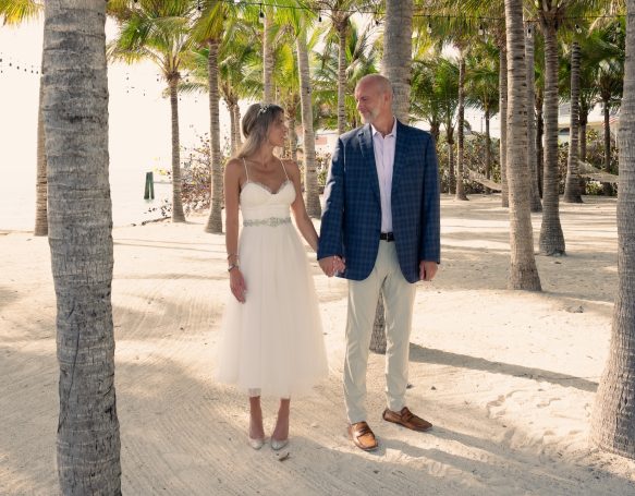 A couple holds hands on a sandy beach, surrounded by palm trees, both smiling.