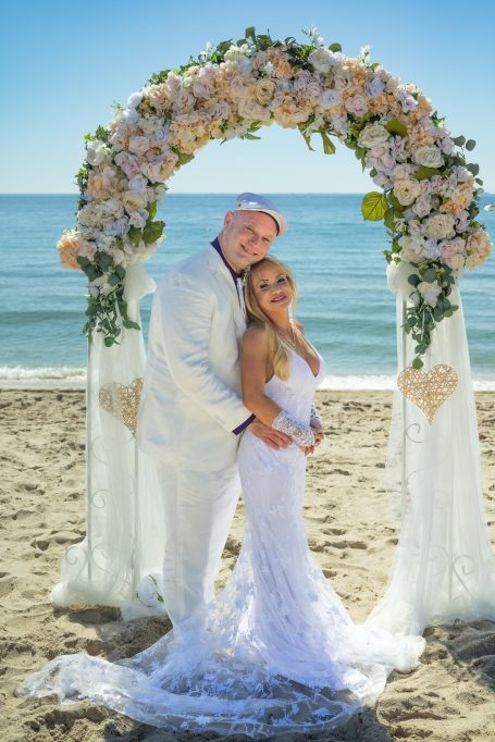 A joyful couple in wedding attire poses under a floral arch on the beach.