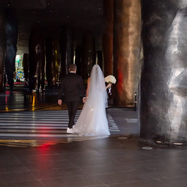 A couple walks hand-in-hand, at The W Hotel, bride in a white gown, beneath tall, dark pillars at The W Hotel