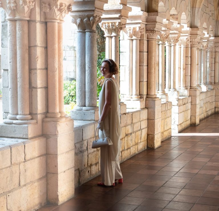 A woman in a white outfit stands by arched stone columns in a sunny corridor.