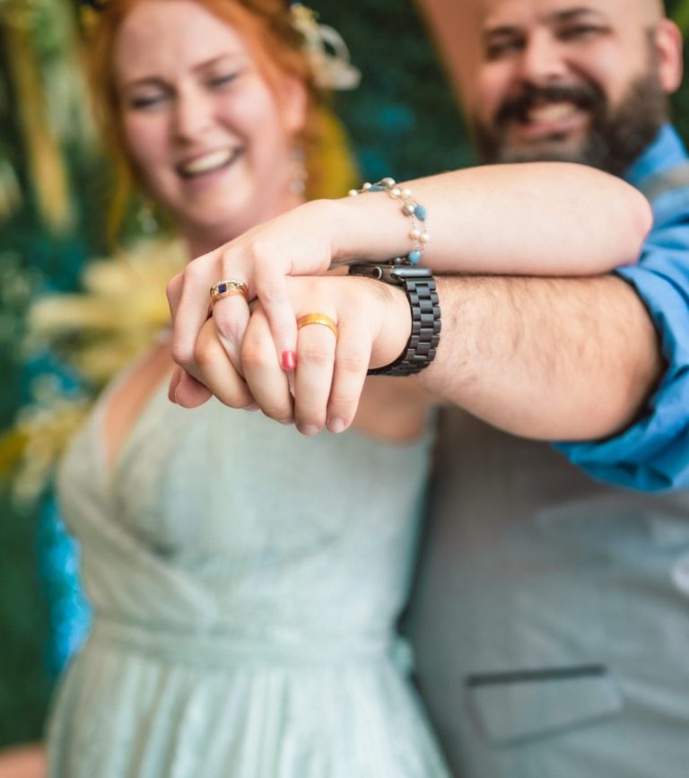 A couple joyfully holding hands, smiling at the camera against a colorful backdrop by Iris Ramil Wedding Officiant.
