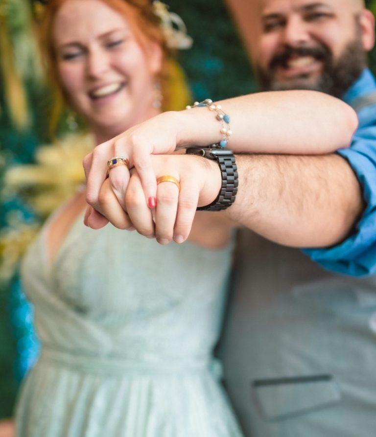 A couple joyfully holding hands, smiling at the camera against a colorful backdrop by Iris Ramil Wedding Officiant.