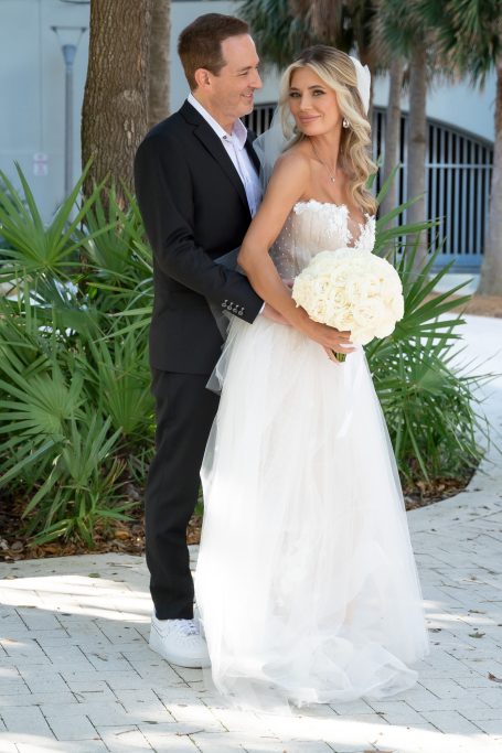 Bride in a white gown holds a bouquet, posing with a groom in a suit outdoors.