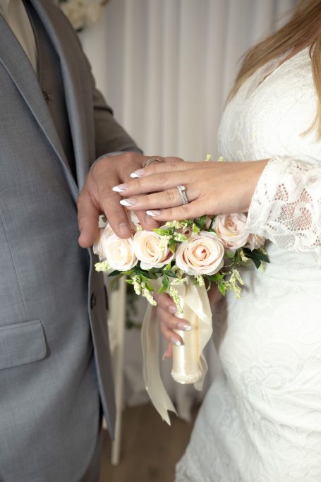 A wedding couple holding hands, showcasing an engagement ring and a bouquet of pink roses.