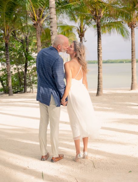Couple kissing on a beach surrounded by palm trees, wearing wedding attire.