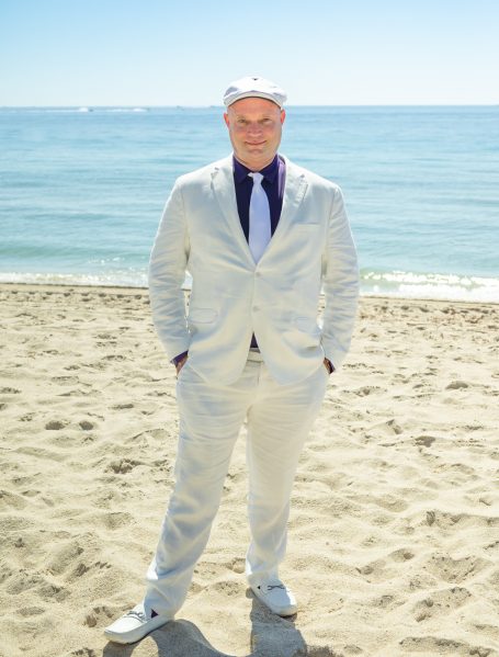 Man in a white suit stands on the beach with a clear blue ocean in the background.