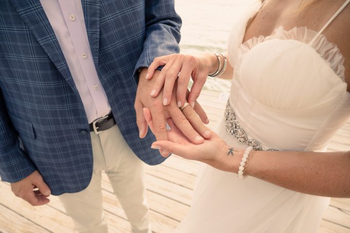 Bride and groom holding hands, showcasing wedding rings on a sunny outdoor setting.