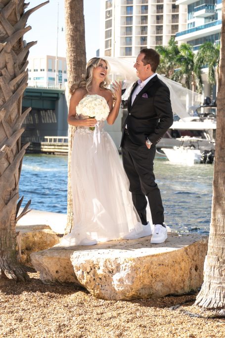 Bride and groom pose together by the water, surrounded by palm trees.