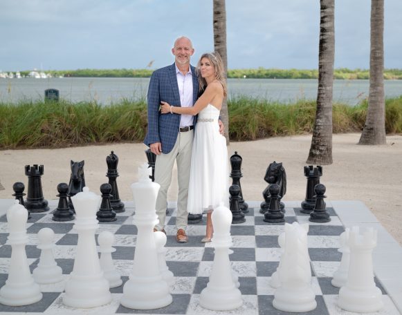 A couple poses together on a giant chessboard near palm trees and a water backdrop.