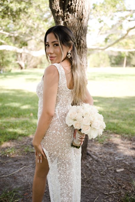 Bride in a sparkling white gown holds a bouquet of white flowers, standing outdoors.