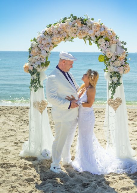 Bride and groom under a floral arch on a beach with ocean views.