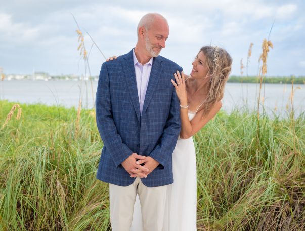 A couple shares a joyful moment by the water, surrounded by tall grass.