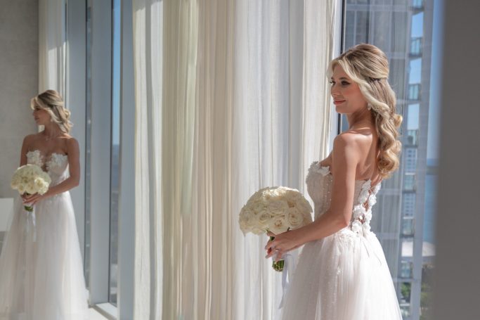 Bride in a white gown holding a bouquet, standing by large windows with sheer curtains.