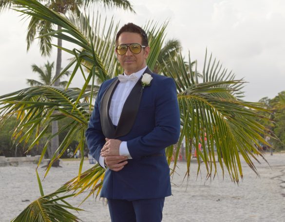 Man in a blue tuxedo with sunglasses, standing near palm trees on a beach.