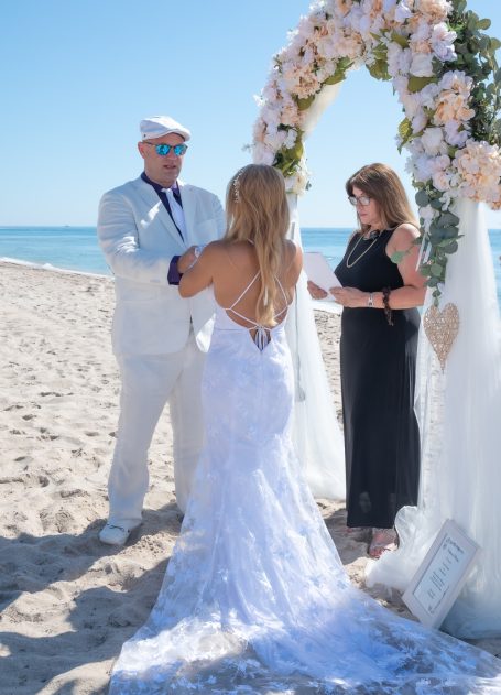 A beach wedding ceremony with a bride in a white dress, groom in white suit, and officiant.
