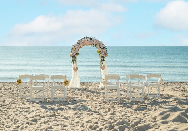Beach wedding setup with an arch of flowers and white chairs facing the ocean.