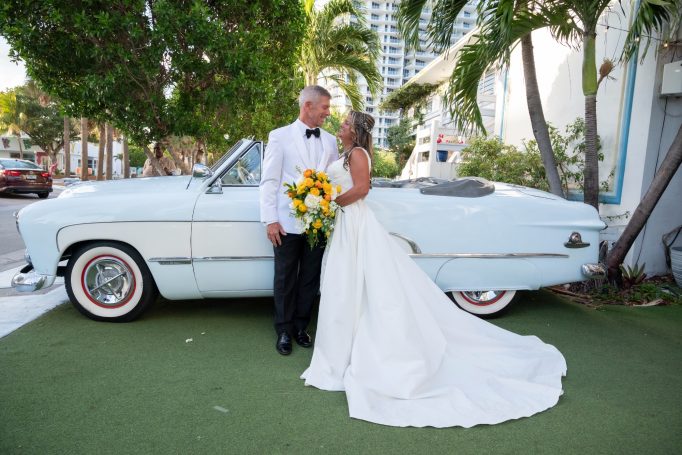 Bride and groom standing beside a vintage white convertible car, surrounded by greenery.