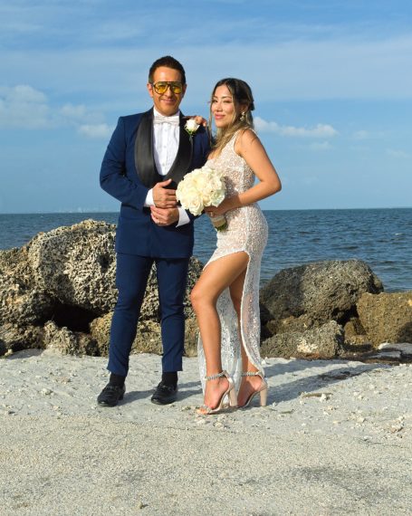 Couple in formal attire poses on a beach, holding flowers with the ocean in the background.