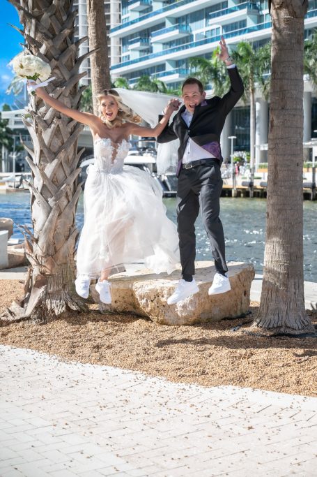 Bride and groom joyfully jumping together near water and palm trees.