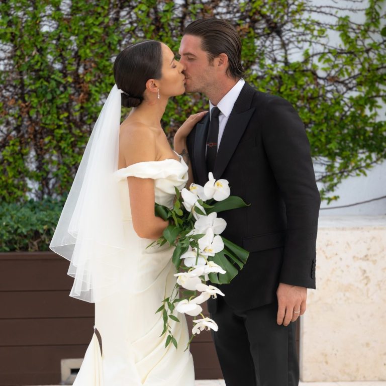Bride and groom share a kiss, surrounded by greenery, with flowers in the bride's hand.