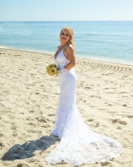 Bride in a white gown holding sunflowers, standing on a sandy beach by the ocean.