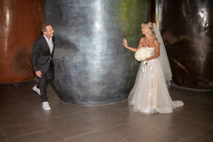 A bride and groom interact playfully near large, colorful metal sculptures indoors.