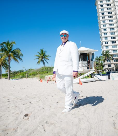 Man in a white suit walking on a beach with palm trees and a lifeguard tower in the background.
