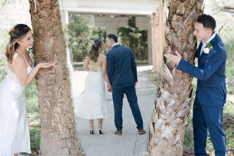 A bride and groom hold hands, framed by two trees, in a sunny outdoor setting.
