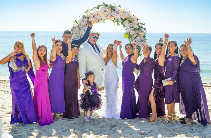 Wedding party in purple attire celebrating on the beach, with a floral arch in the background.