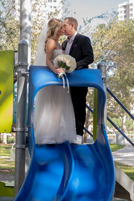 Bride and groom kiss on a playground slide, surrounded by greenery.