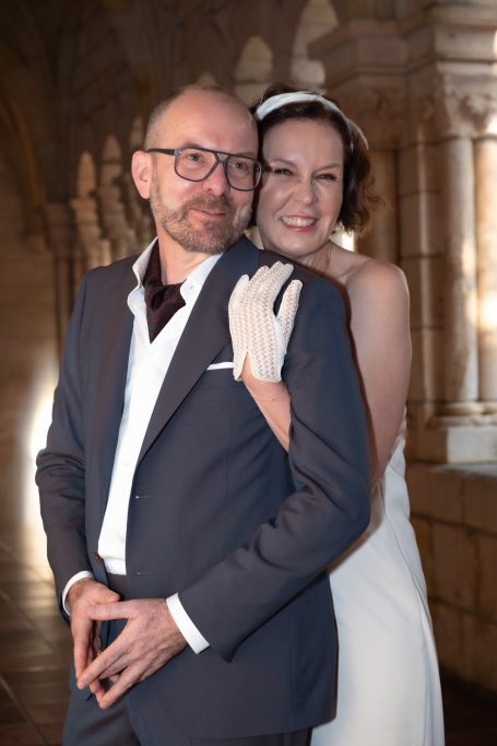 A smiling couple in formal attire, with the woman wearing a wedding dress and gloves.