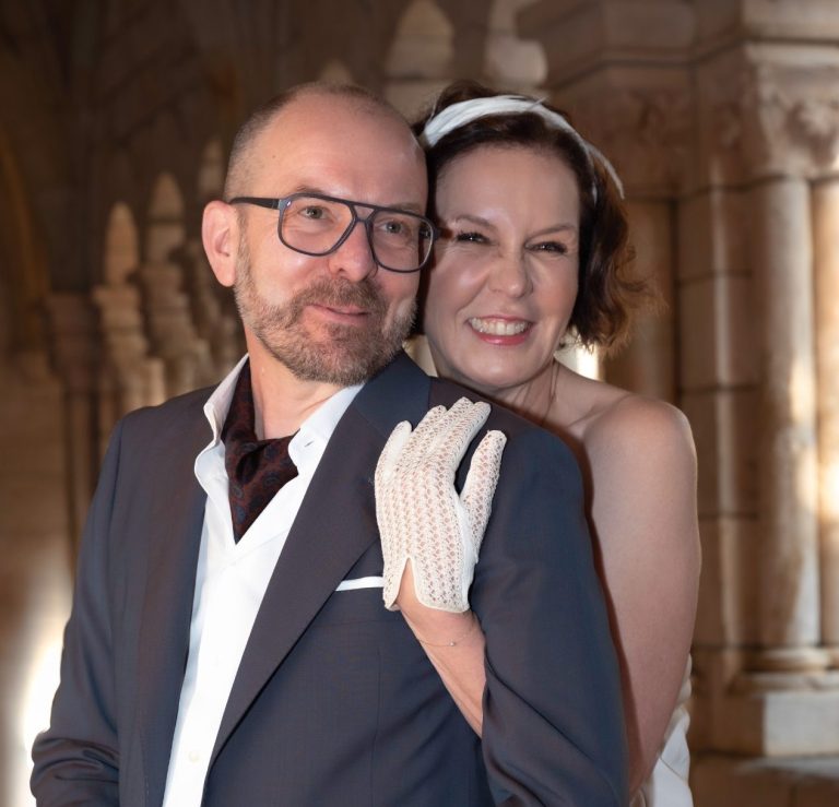 A smiling couple poses together, dressed in formal attire in a beautiful location.
