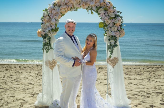 Bride and groom posing under a floral arch on the beach with ocean in the background.