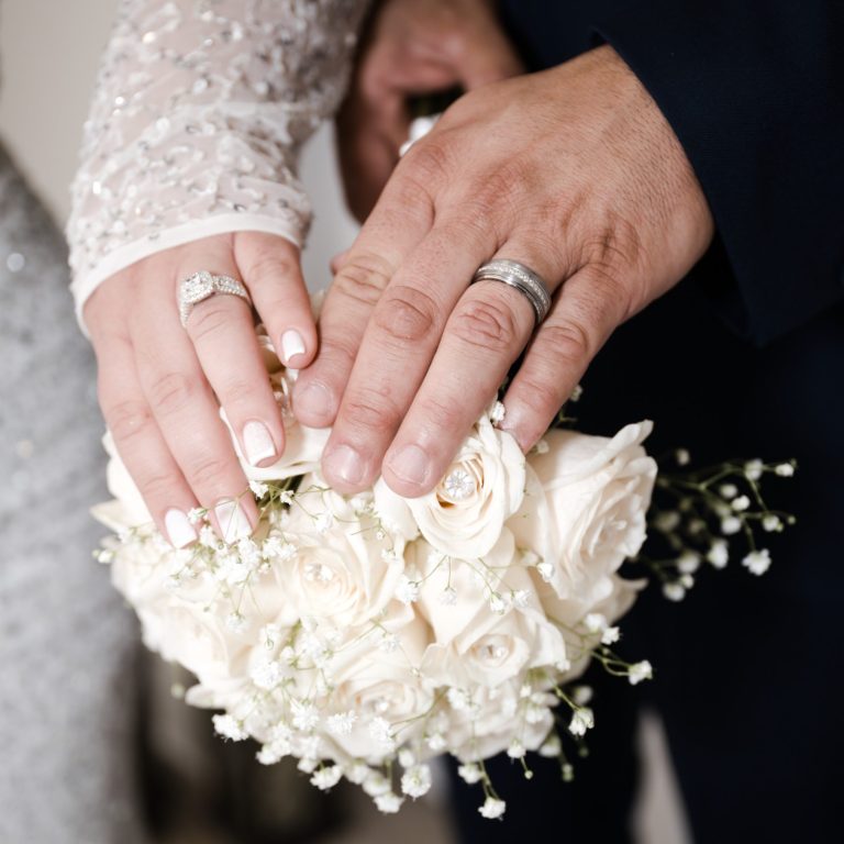 Close-up of bride & groom's hands with wedding rings holding a bouquet of white roses & baby’s breath.