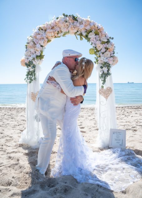 Couple embracing under a floral arch on a sunny beach.