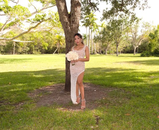 Woman in a flowing dress holding a bouquet, standing in a green park under a tree.