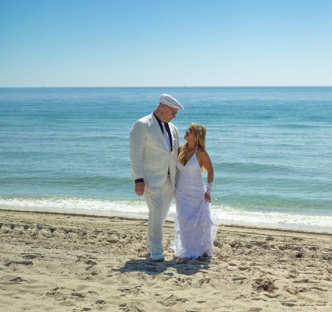 A couple in wedding attire stands on a sandy beach by the calm ocean.