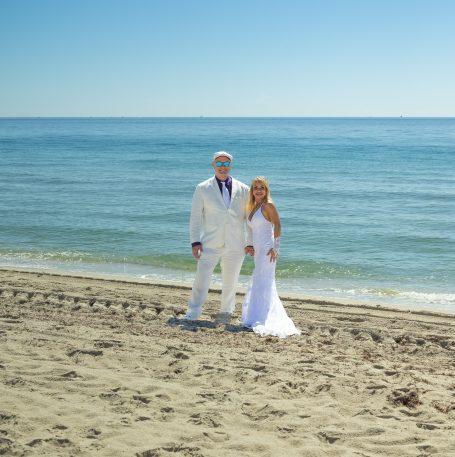 A couple in formal wedding attire stands on a sandy beach by the ocean.
