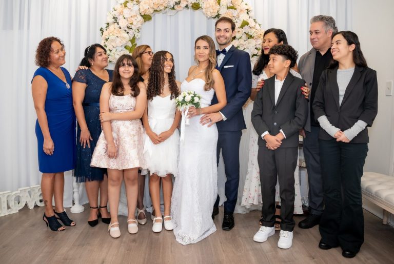 A bride and groom hold hands, framed by two trees, in a sunny outdoor setting.