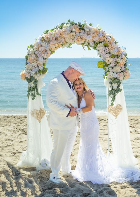 A bride and groom stand under a floral arch on the beach, smiling happily at each other.